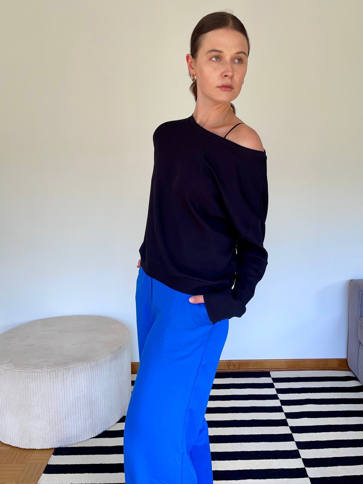 Woman wearing a black off-shoulder Pia top and Bright blue Han pants standing in a room with a checkered rug.