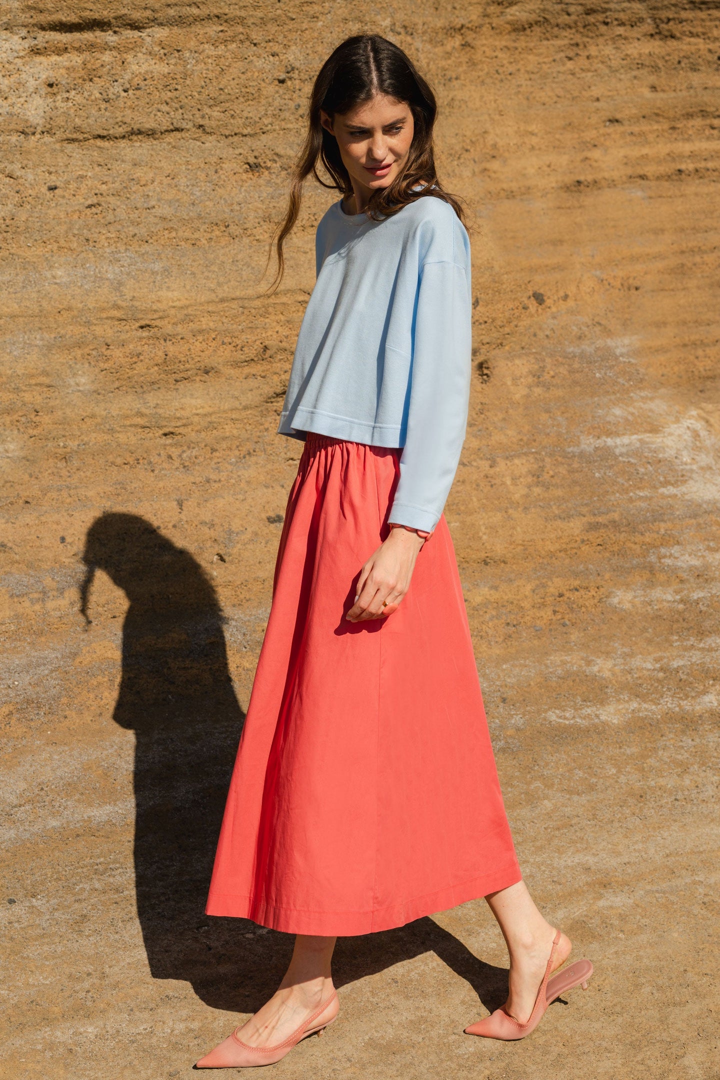 Woman in a light blue top and pink skirt standing against a textured brown wall.
