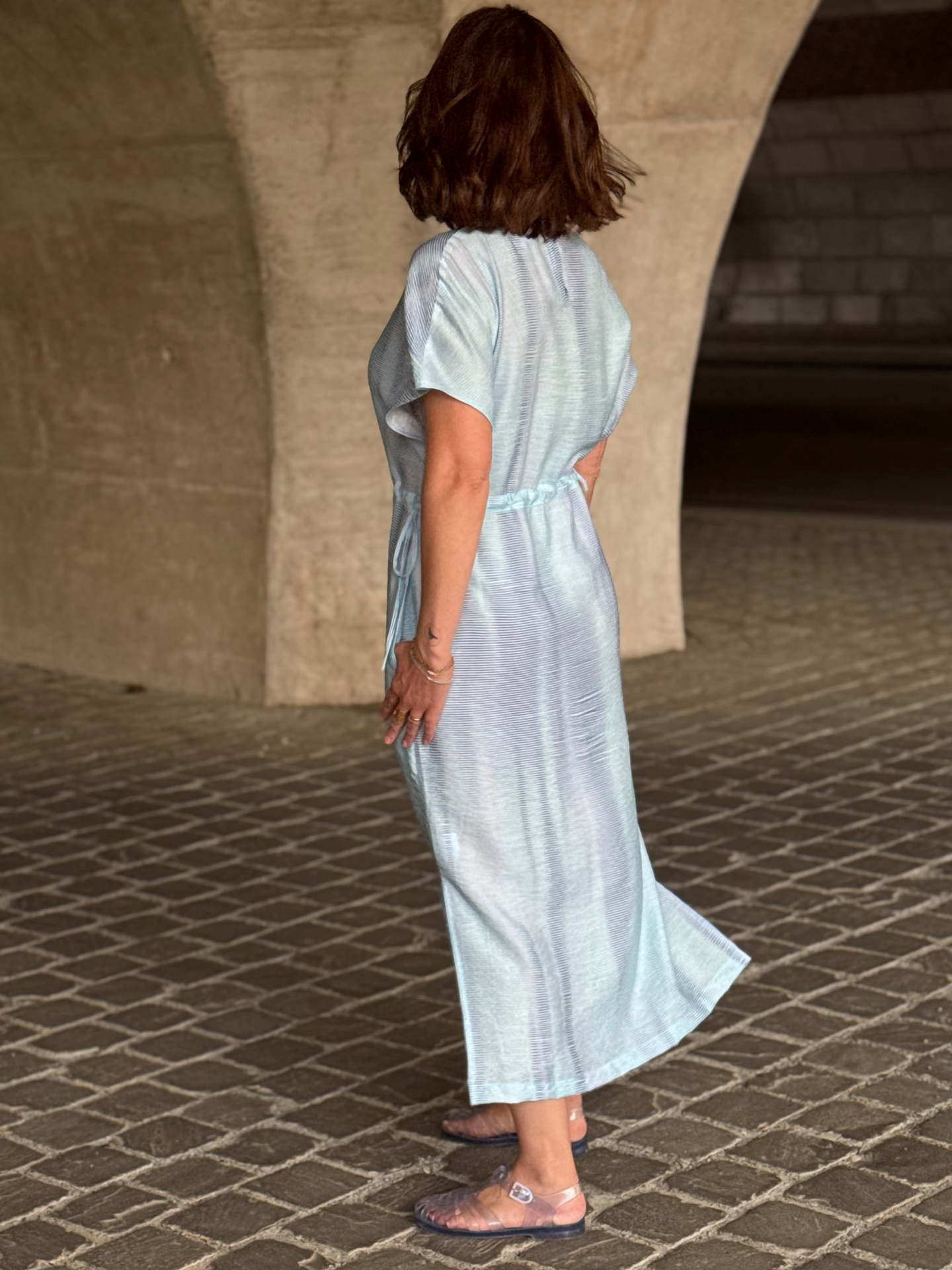 Woman in a light blue dress standing on a stone pavement with a concrete wall in the background.