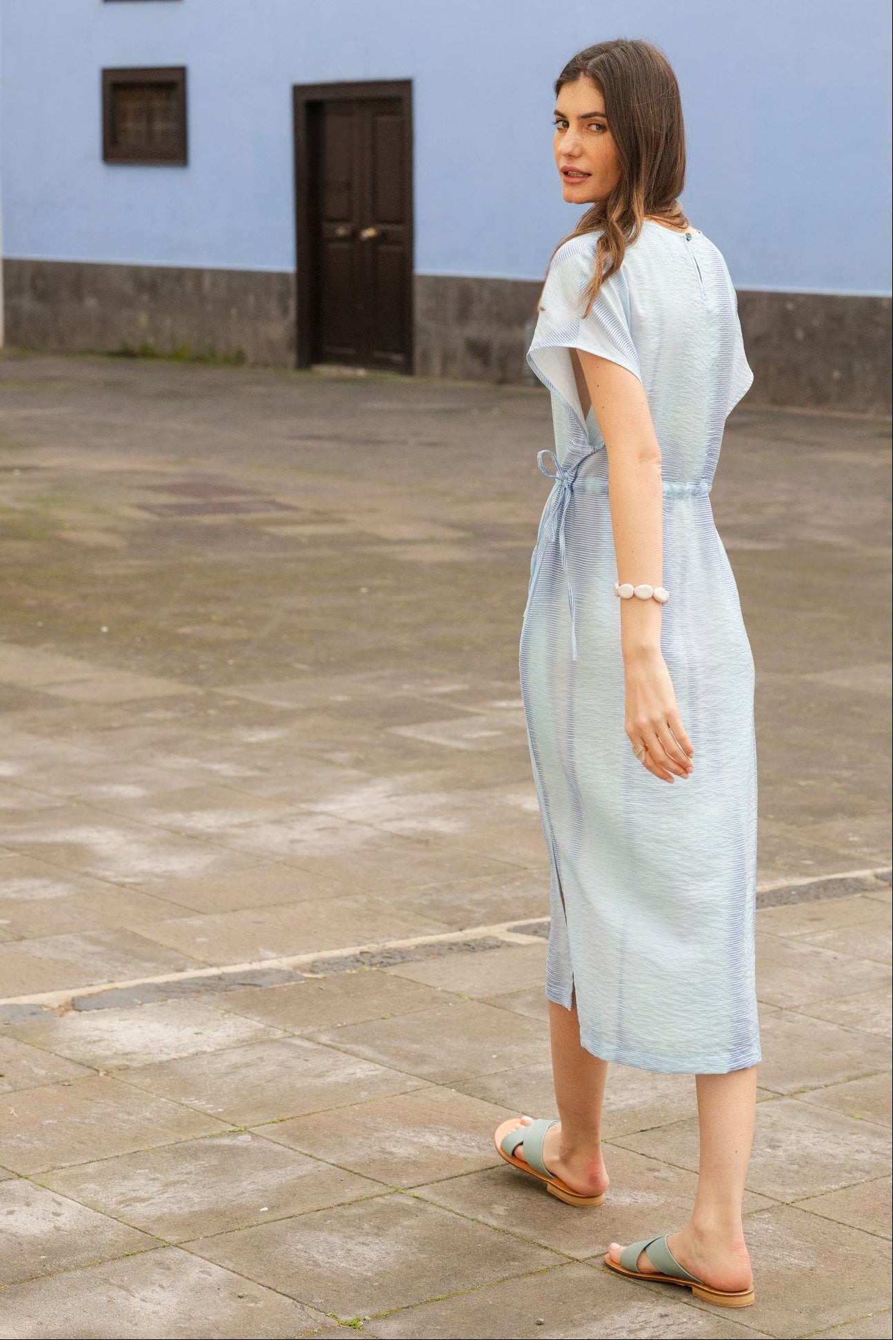 Woman in a light blue dress standing in front of a blue wall with windows.