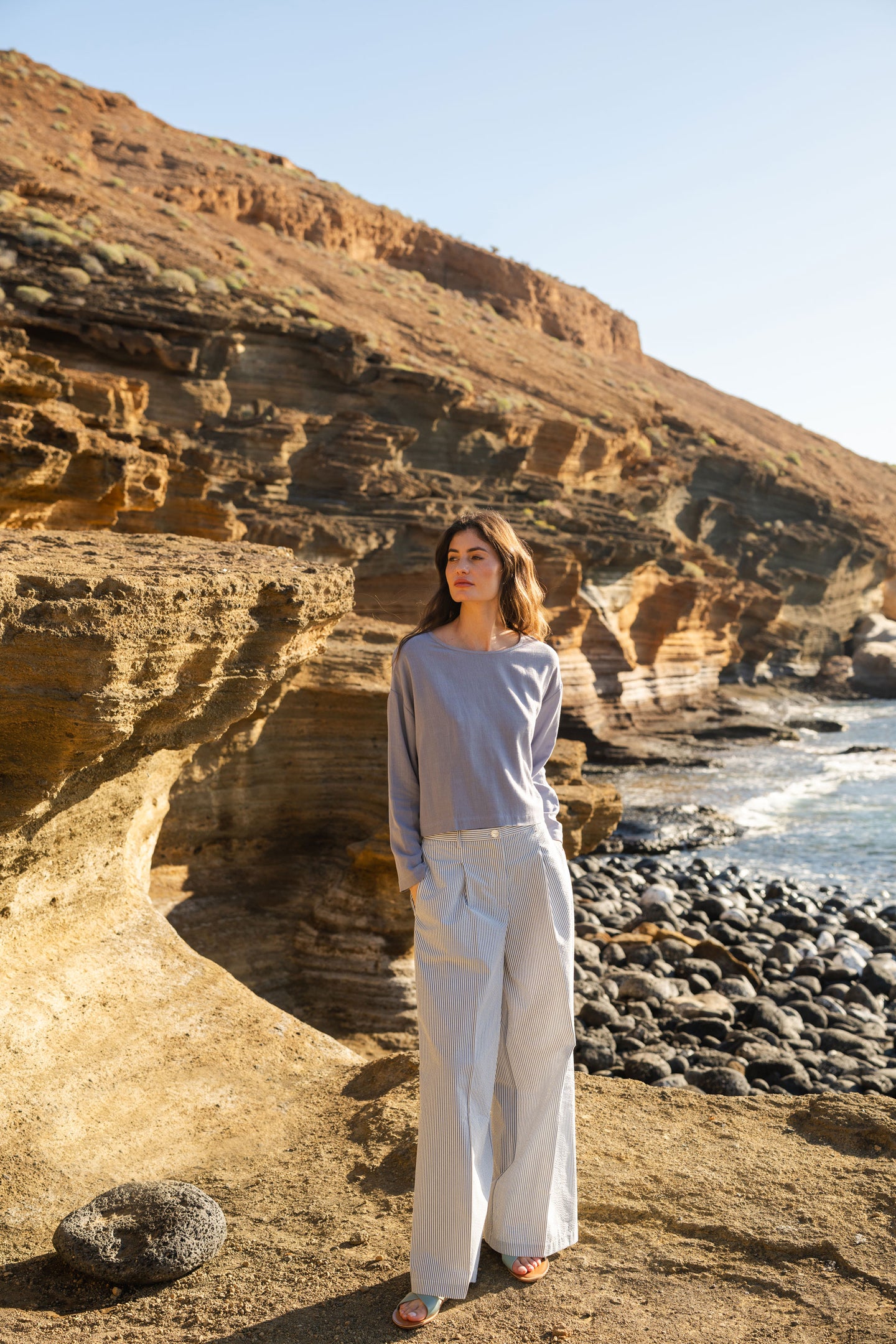 Woman standing on a rocky beach with cliffs and ocean in the background in a blue stone blouse and striped seersucker pants.