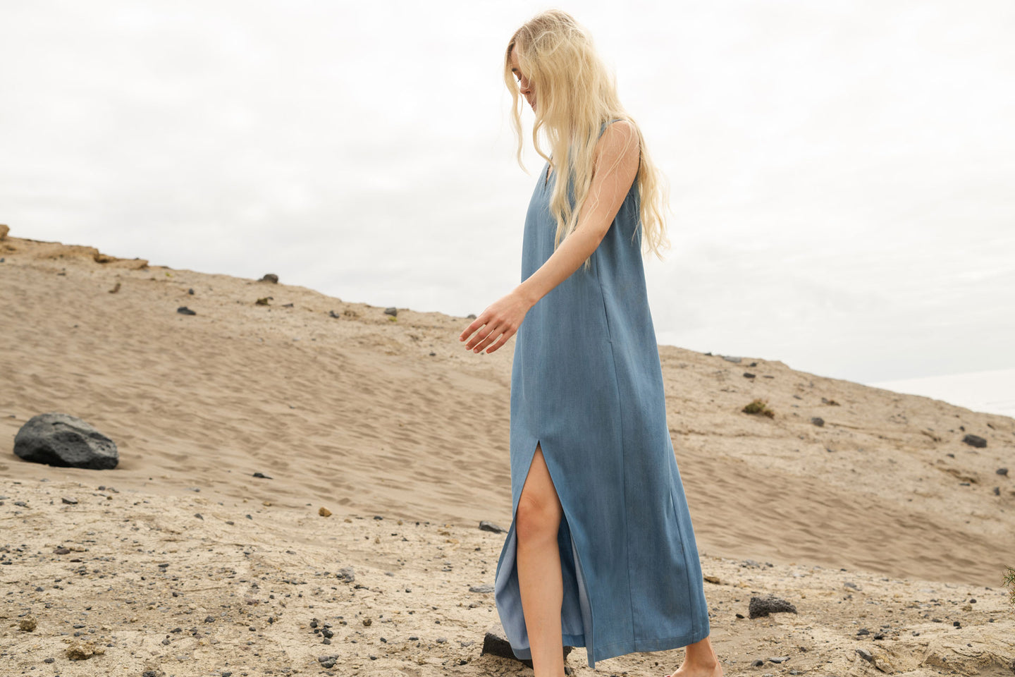 Woman in a light blue dress walking on a stone pavement with a blue wall and windows in the background.