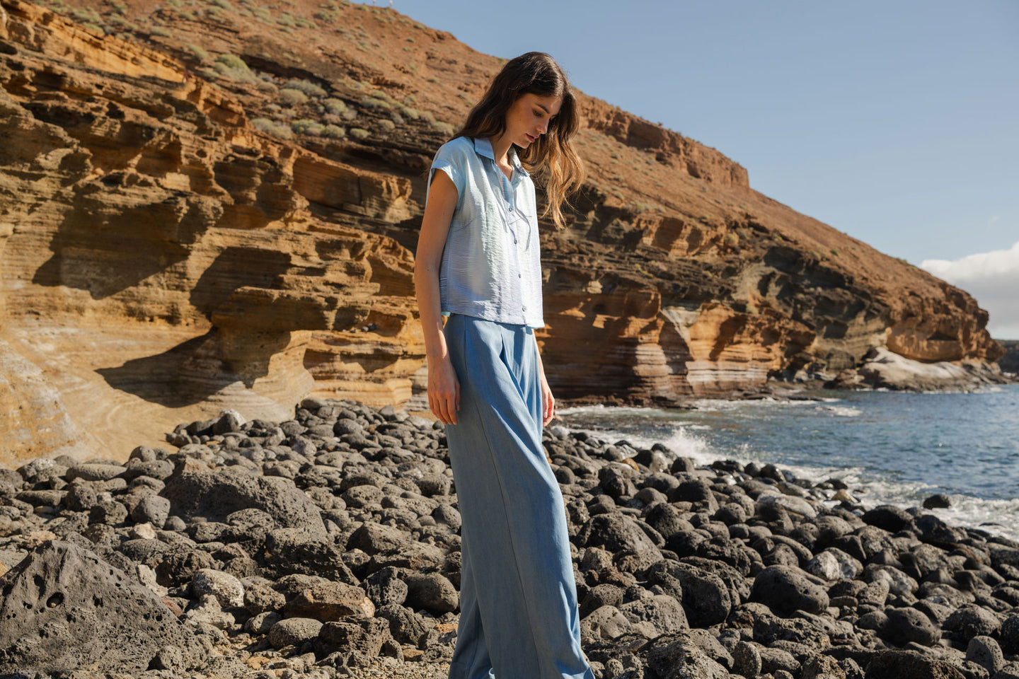Woman standing on a rocky beach with cliffs and ocean in the background in a light blue blouse & jeans.