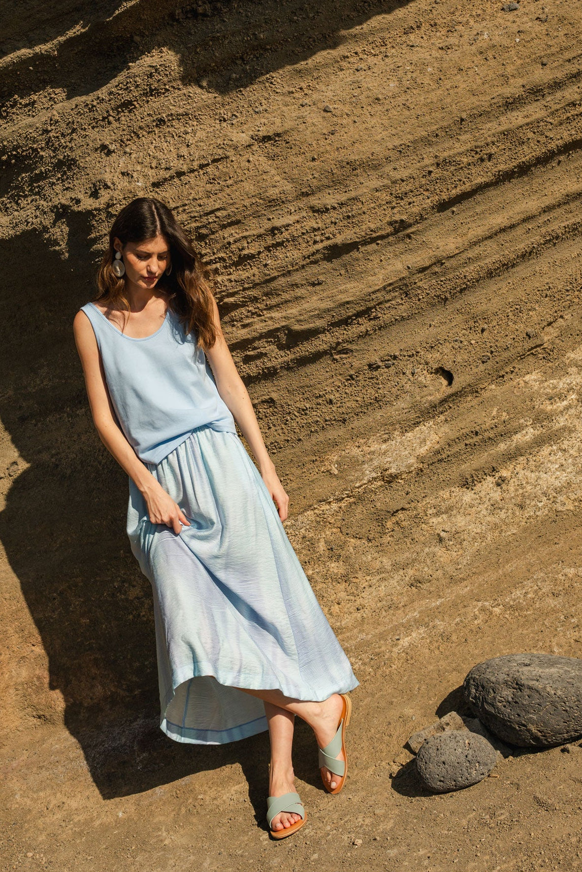 Woman in a light blue top & skirt standing on a rocky, sunlit landscape.
