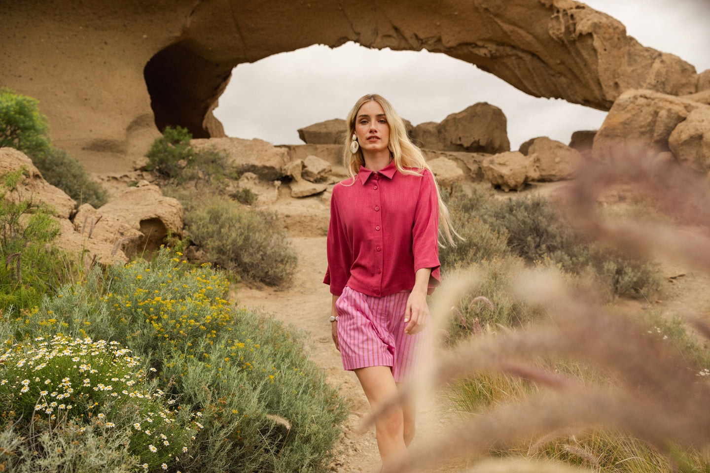 Woman in a magenta shirt and pink striped shorts standing under a natural arch in a desert landscape.