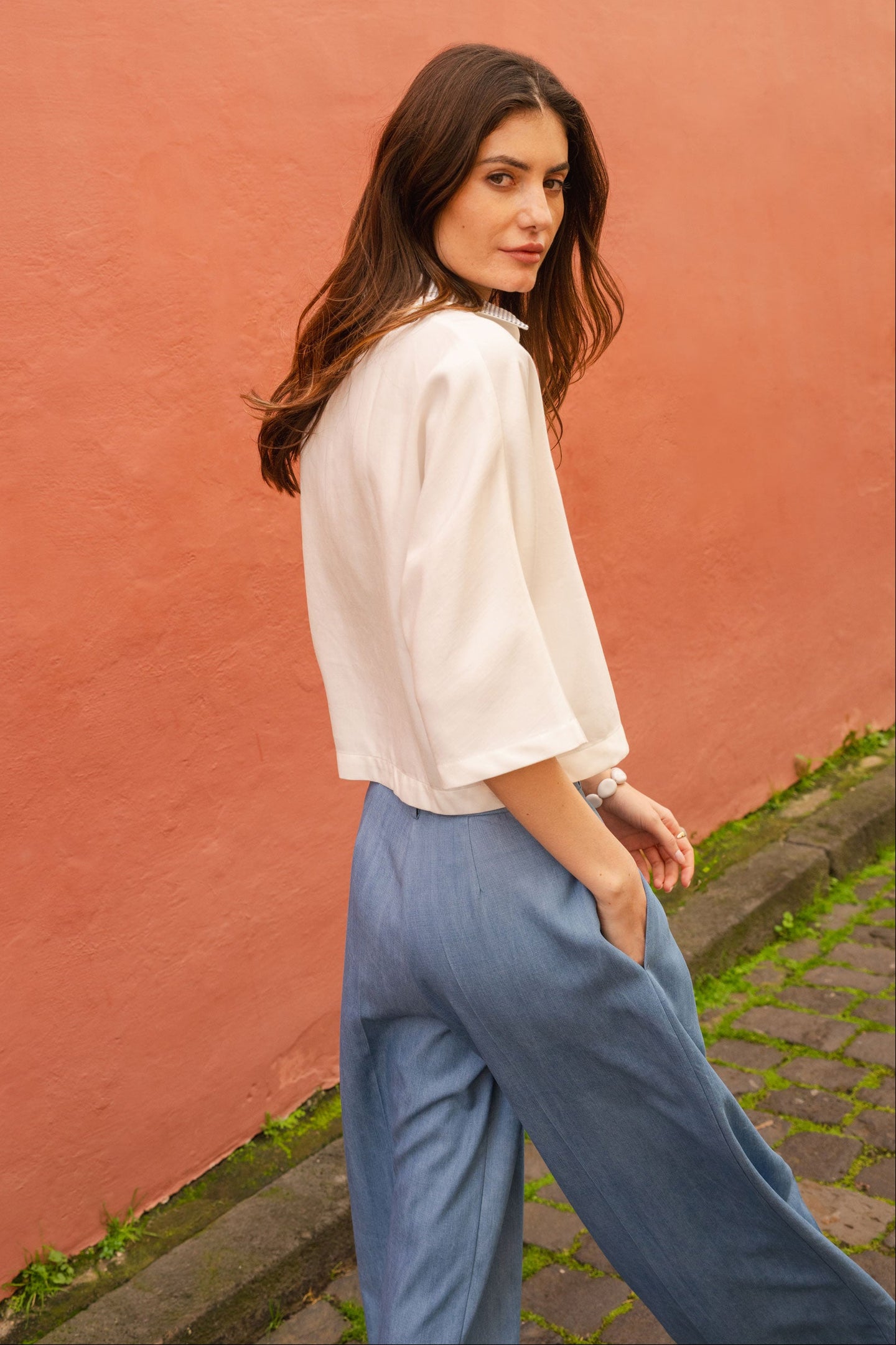 Woman in a white blouse and blue jeans pants standing against a pink wall.