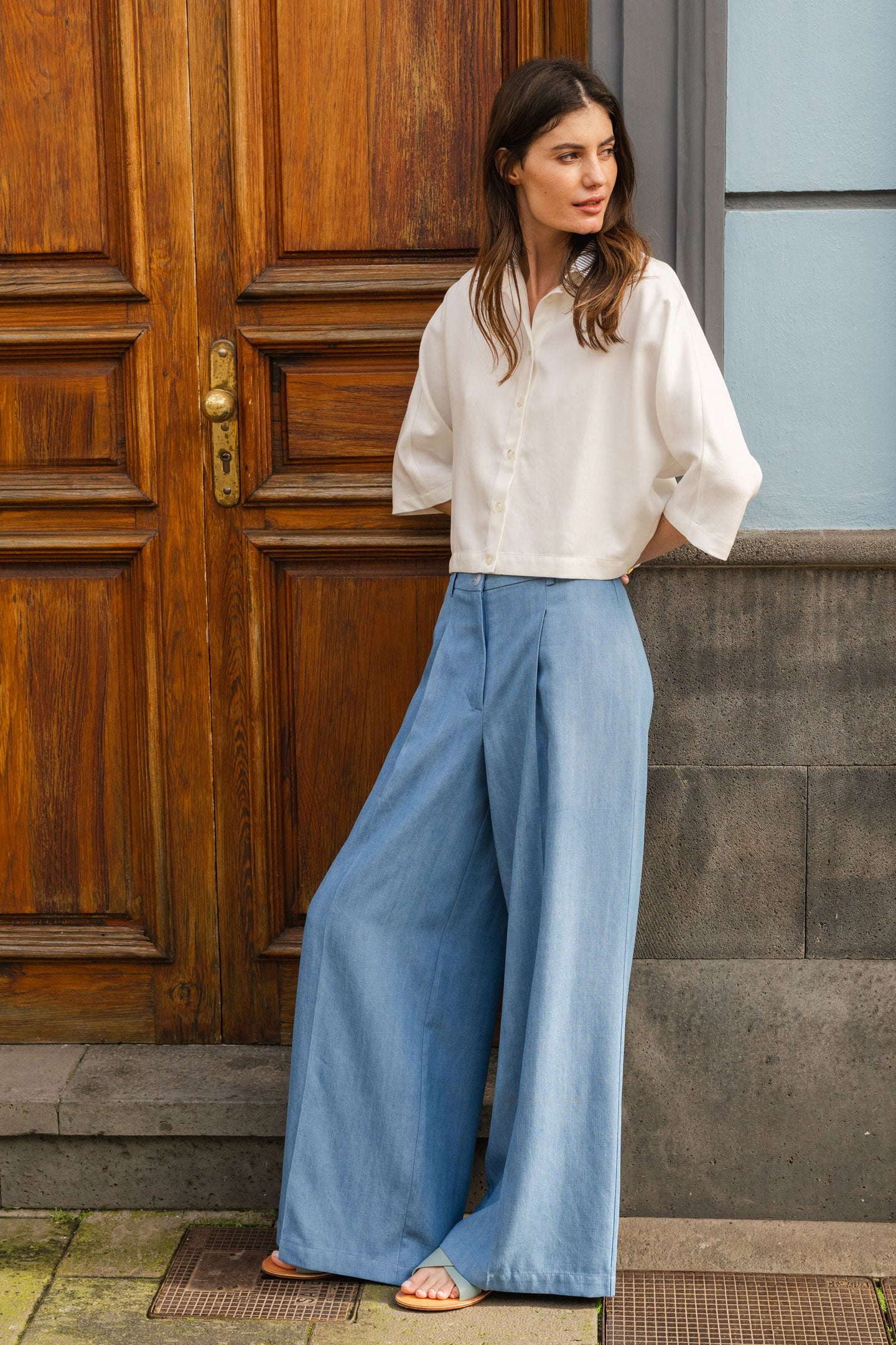 Woman wearing a white blouse and blue jeans pants standing in front of a wooden door.