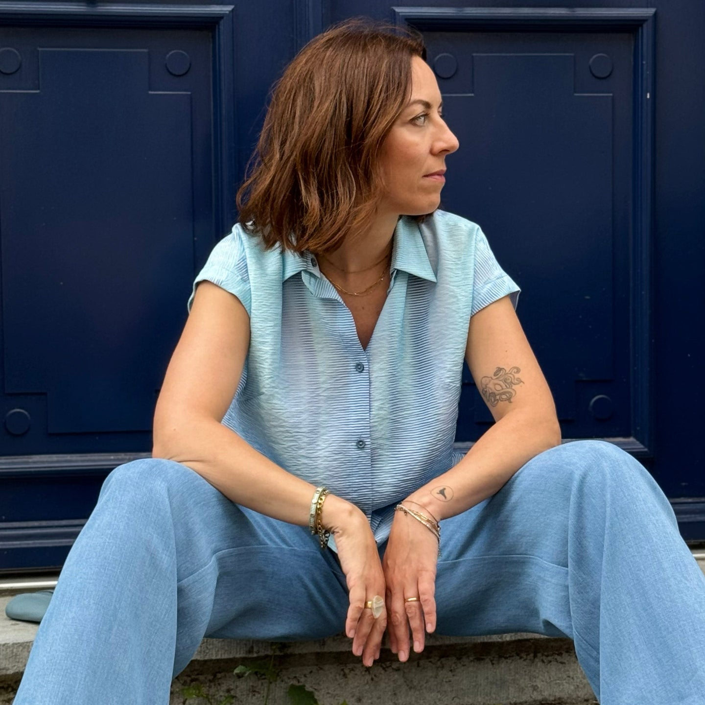 Woman in a light blue blouse & jeans sitting against a blue door.