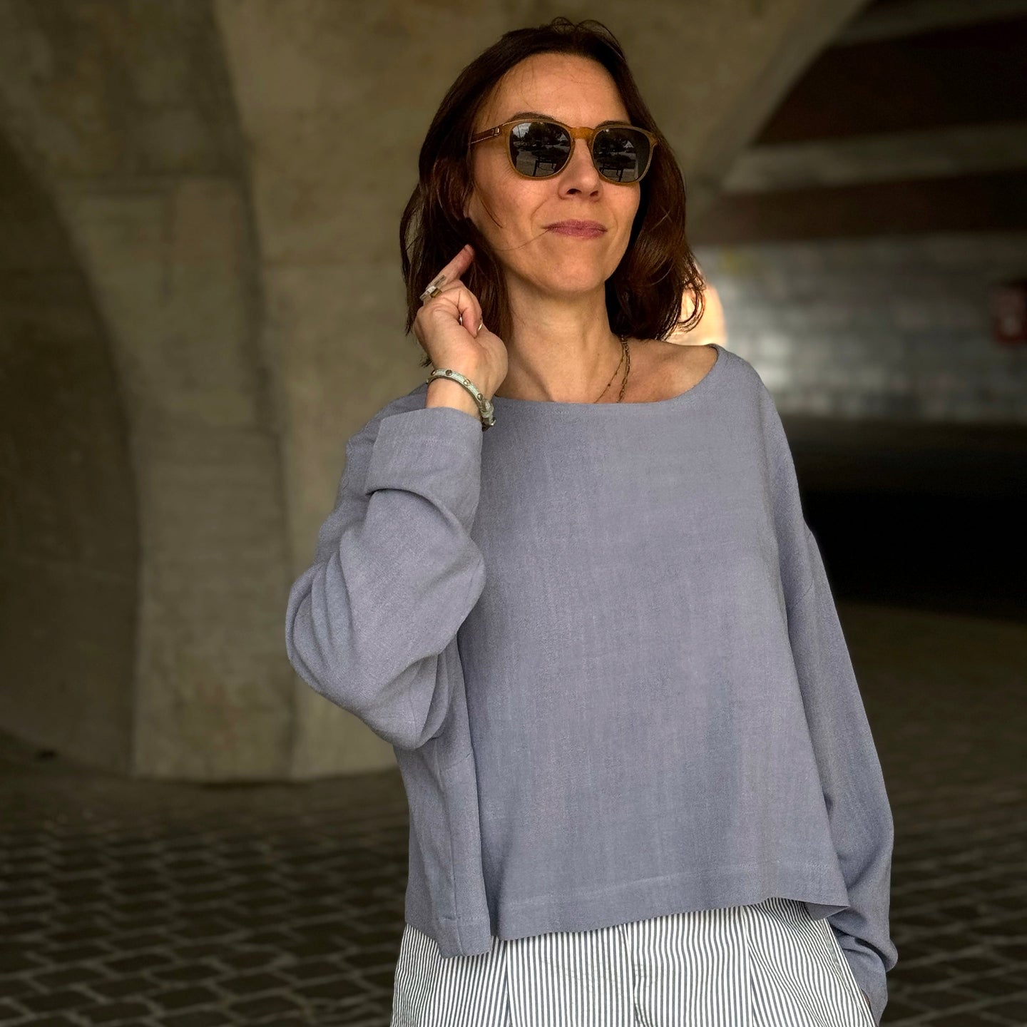 Woman wearing sunglasses and a blue stone top standing under a stone archway.