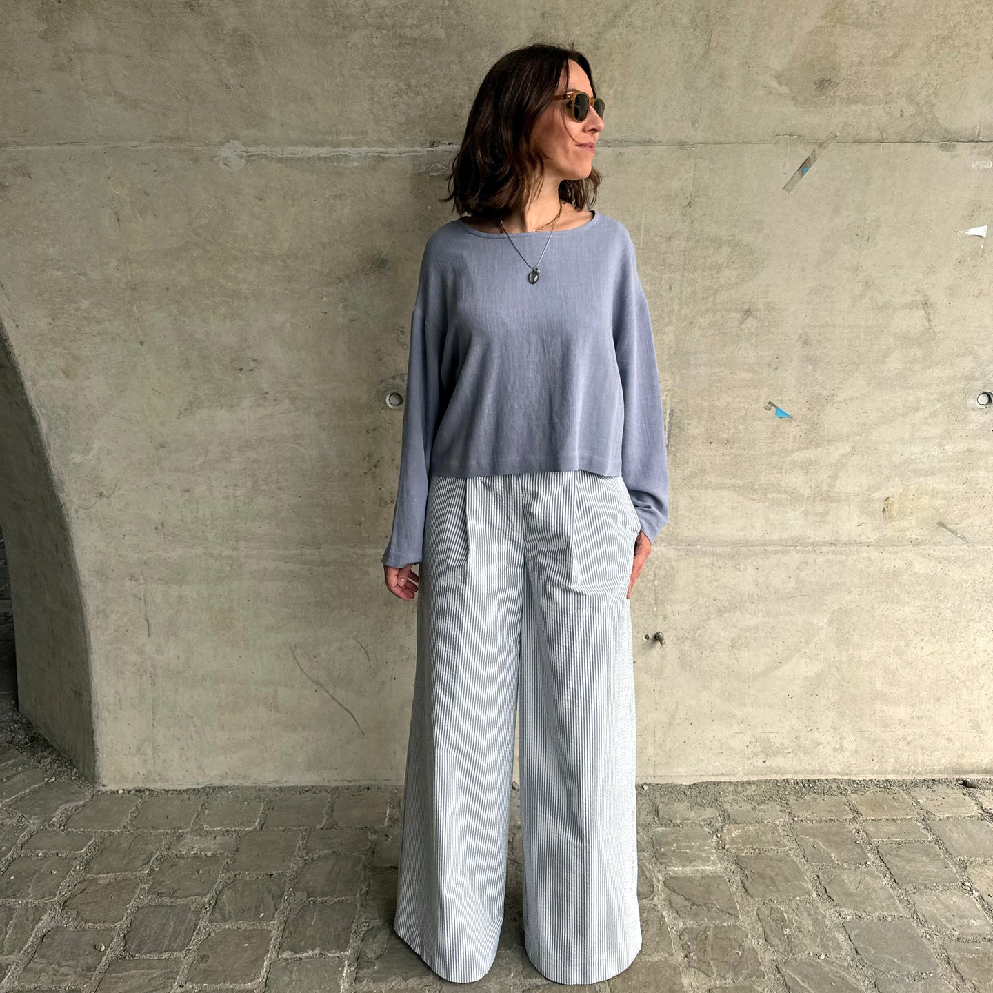 Person wearing a light blue top and striped pants standing against a concrete wall.