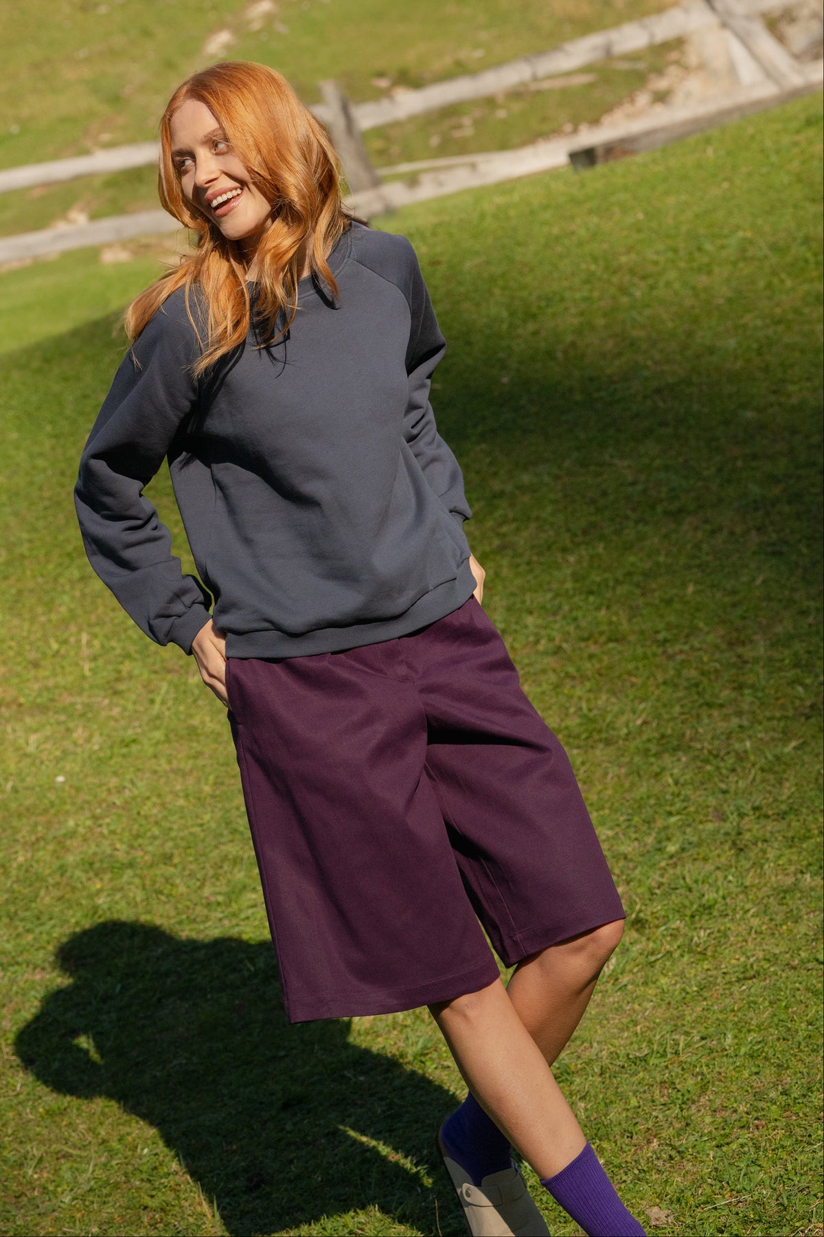 A woman with red hair stands on a grassy hill wearing a dark gray sweater and a dark purple knee-length skirt, with trees and a wooden fence in the background.