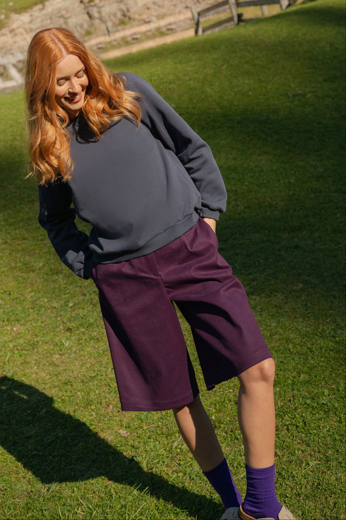 A woman with long red hair stands outdoors on a grassy hill wearing a dark gray sweater, purple knee-length shorts, and purple socks, with trees and a rocky slope in the background.