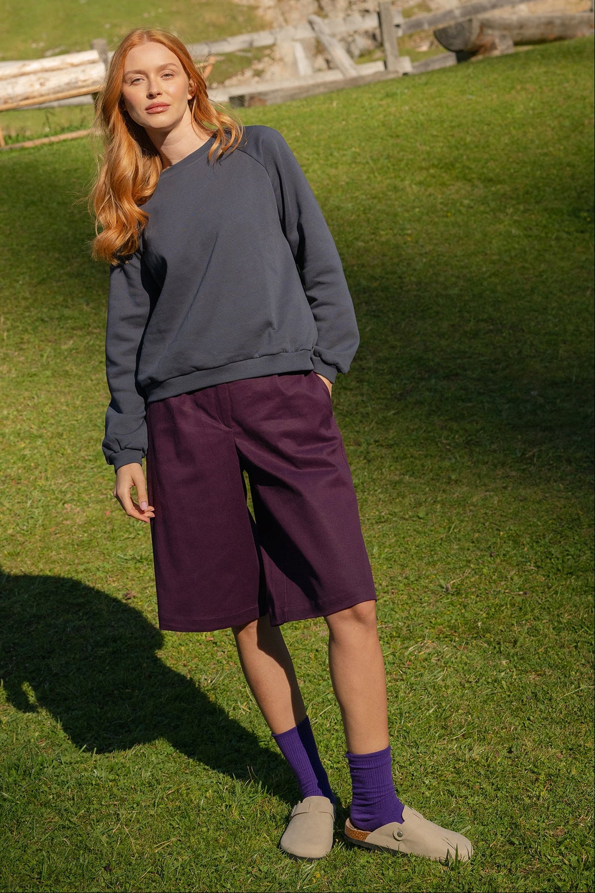 A woman stands on a grassy mountain path wearing a meteorite sweater, purple knee-length shorts, purple socks, and beige slip-on shoes, with trees and a wooden fence in the background.